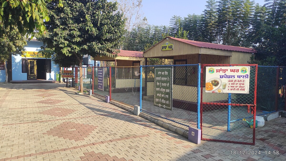A fenced area is visible, featuring two wooden structures with gabled roofs. Signage is displayed in multiple languages, and the paved ground is surrounded by trees. A clear pathway leads toward the entrance of the facility, creating an accessible outdoor environment.