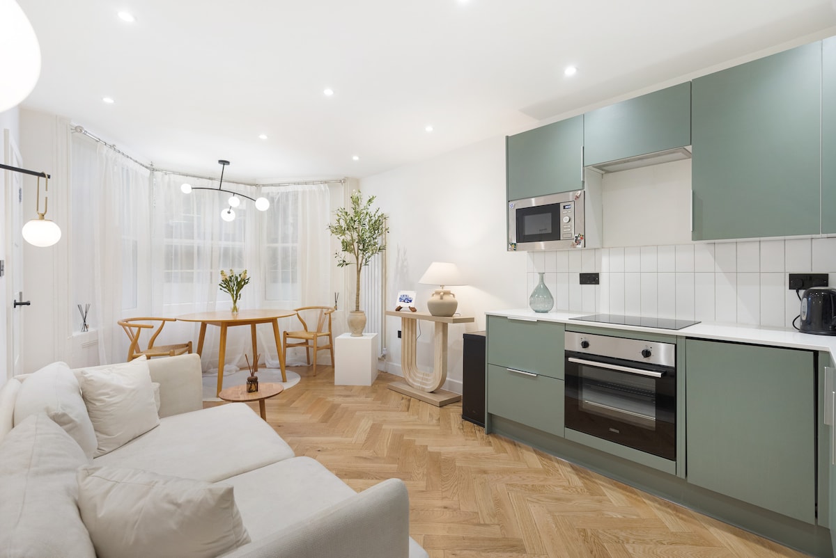 The modern kitchen features mint green cabinetry and stainless-steel appliances, including an oven and microwave. A beige sofa sits in the living area, complemented by a dining table with wooden chairs. Natural light enters through sheer curtains, enhancing the space's bright ambiance.