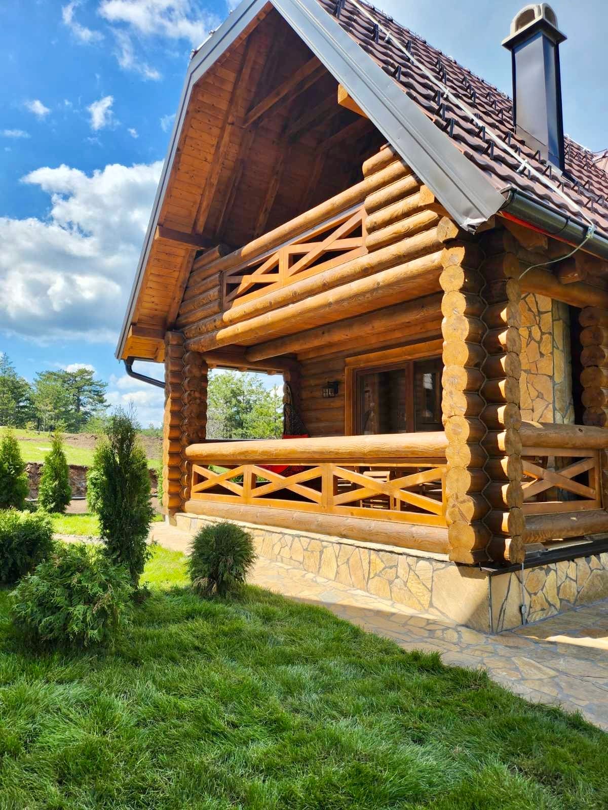 A charming cabin constructed from natural wood features a traditional log design. The front porch showcases decorative railings and offers outdoor seating. Lush green grass and small evergreen shrubs surround the cabin, complemented by a backdrop of trees and an expansive blue sky with scattered clouds.