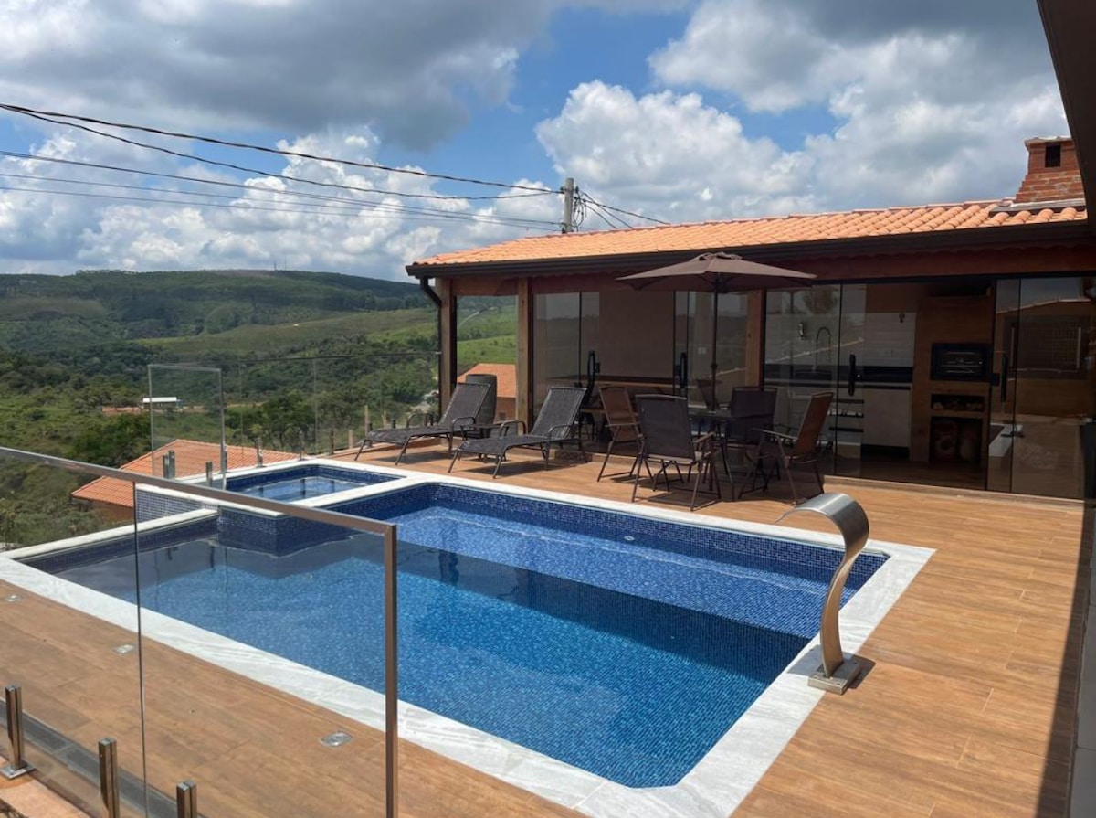 An outdoor terrace features a spacious swimming pool with a built-in ladder and a vibrant blue surface. Surrounding the pool are multiple lounge chairs and an umbrella for shade. A barbecue area is also visible, complemented by scenic views of the surrounding mountains.