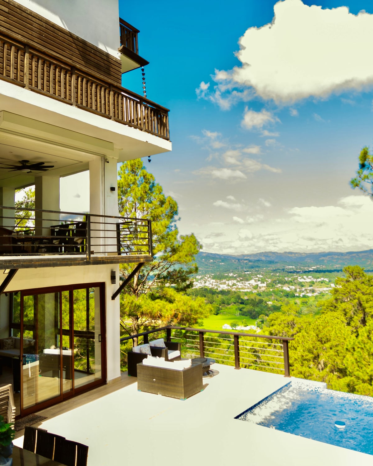 An outdoor area is presented with a private pool overlooking expansive green valleys and mountains. Comfortable seating is arranged on a sunlit patio, and large glass doors connect the interior to the serene outside views, inviting relaxation in a natural setting.
