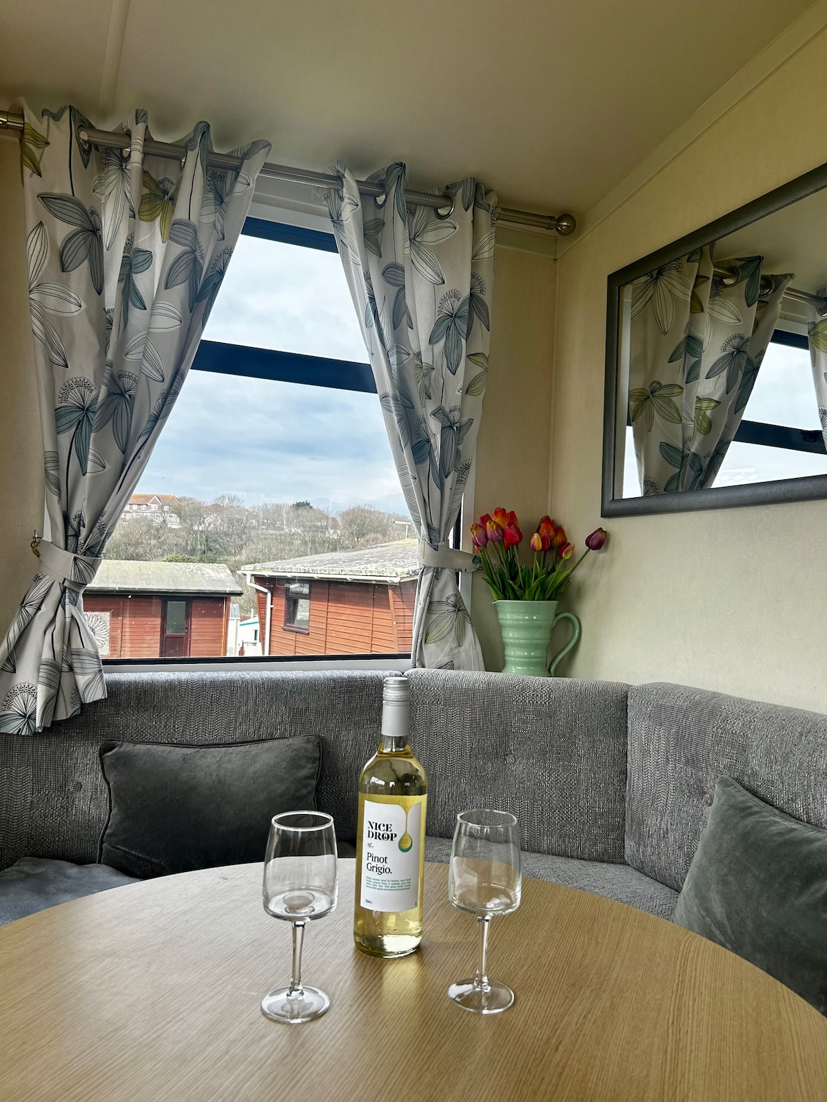 A cozy seating area features a grey upholstered sofa surrounding a circular wooden table. A bottle of white wine and two glasses are placed on the table. Bright curtains with a leaf pattern adorn the window, allowing natural light to illuminate the space.