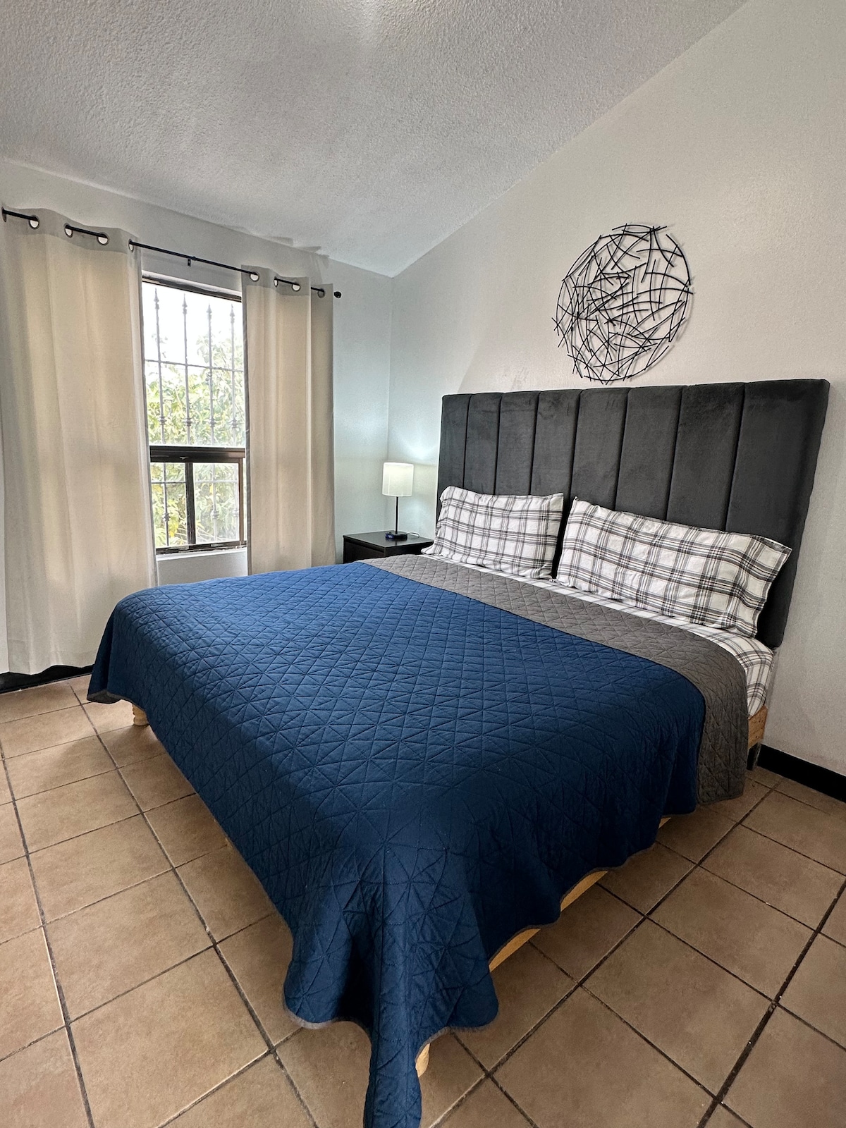 A well-lit bedroom is presented, featuring a spacious bed with a navy coverlet and plaid accent pillows. Large windows dressed with light curtains allow natural light to fill the room. A small bedside table with a lamp enhances the functionality of the space.
