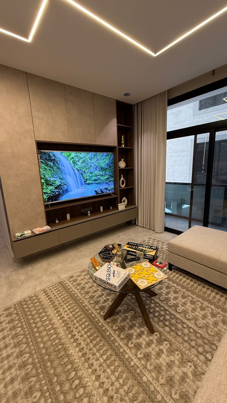 A modern living area is featured, with a large wall-mounted television displaying a nature scene. A light-colored sofa is paired with a round wooden coffee table, showcasing various board games. Floor-to-ceiling windows allow for natural light, and soft curtains frame the view.