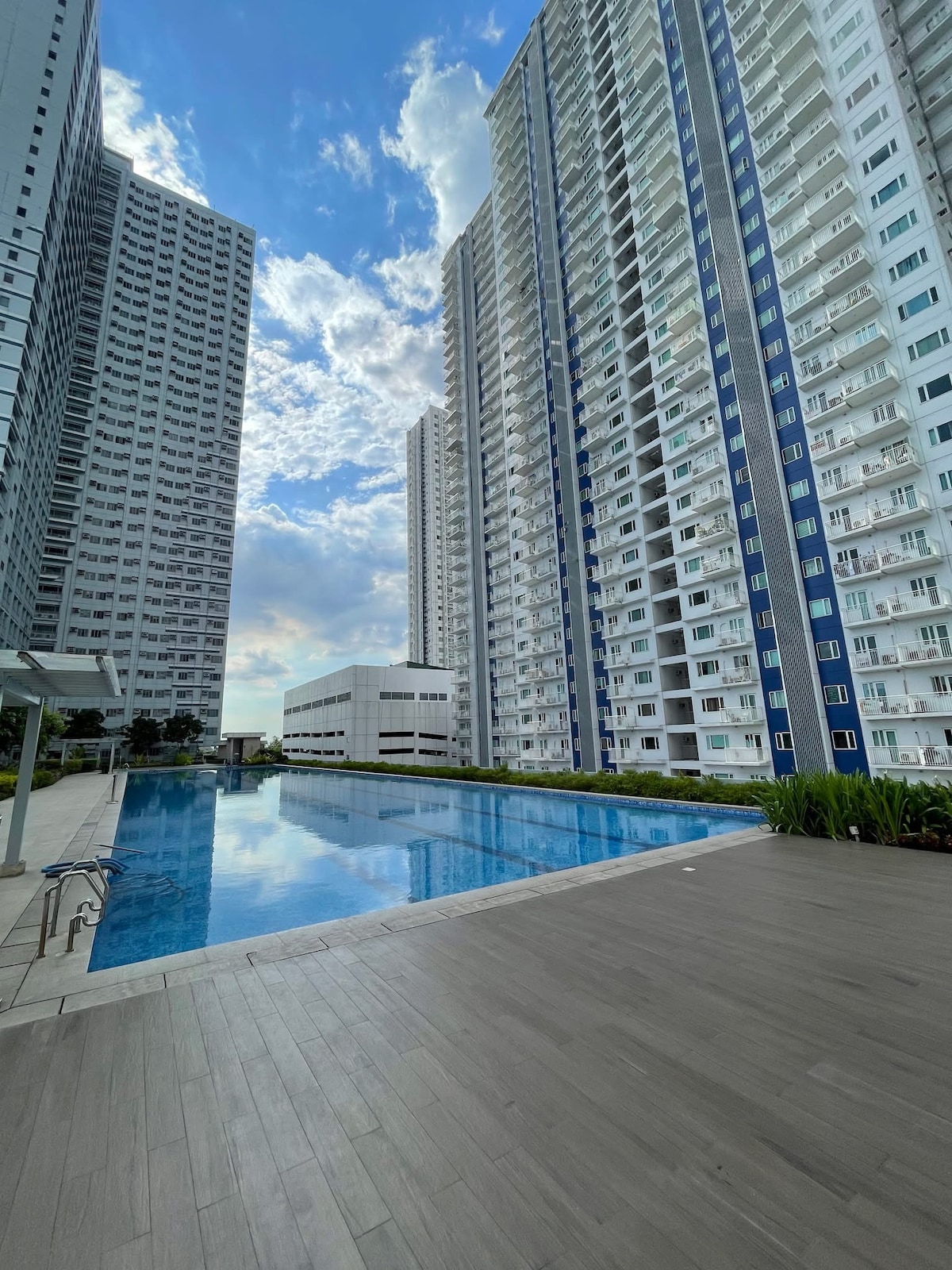 A large swimming pool is visible, surrounded by well-maintained greenery and modern residential towers. The pool reflects the clear blue sky and nearby buildings, creating a serene atmosphere. A poolside area with wooden decking is present, offering space for relaxation and enjoyment.