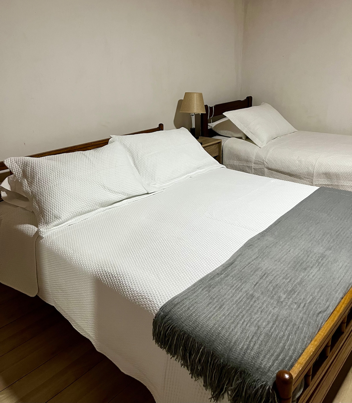 The sleeping area includes two beds, one spacious and one slightly smaller, both dressed in white bedding. A gray throw blanket is draped over the larger bed. A bedside lamp sits on a small table next to the larger bed, contributing to the functionality of the space.