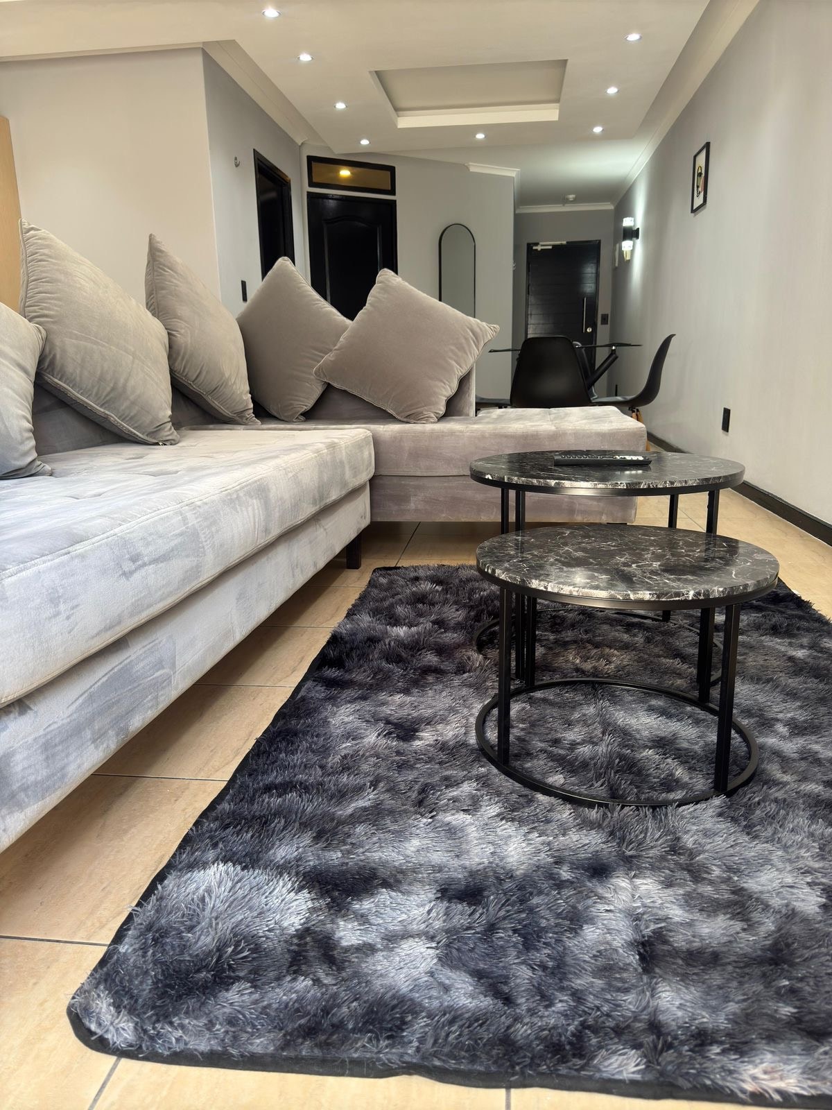 A cozy living area features a plush corner sofa with multiple decorative cushions, complemented by two stylish marble-top coffee tables. A soft, textured rug in shades of grey provides warmth underfoot, while ambient lighting from the ceiling enhances the inviting feel of the space.