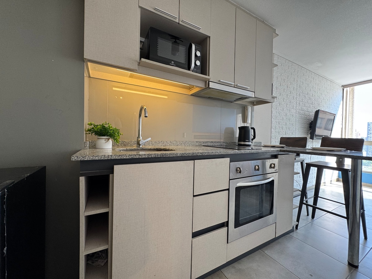 A modern kitchen is equipped with sleek cabinetry and a granite countertop. Essential appliances, including a microwave and coffee maker, are situated on the countertop, while a small plant adds a touch of greenery. A high table and stools provide a space for casual dining.