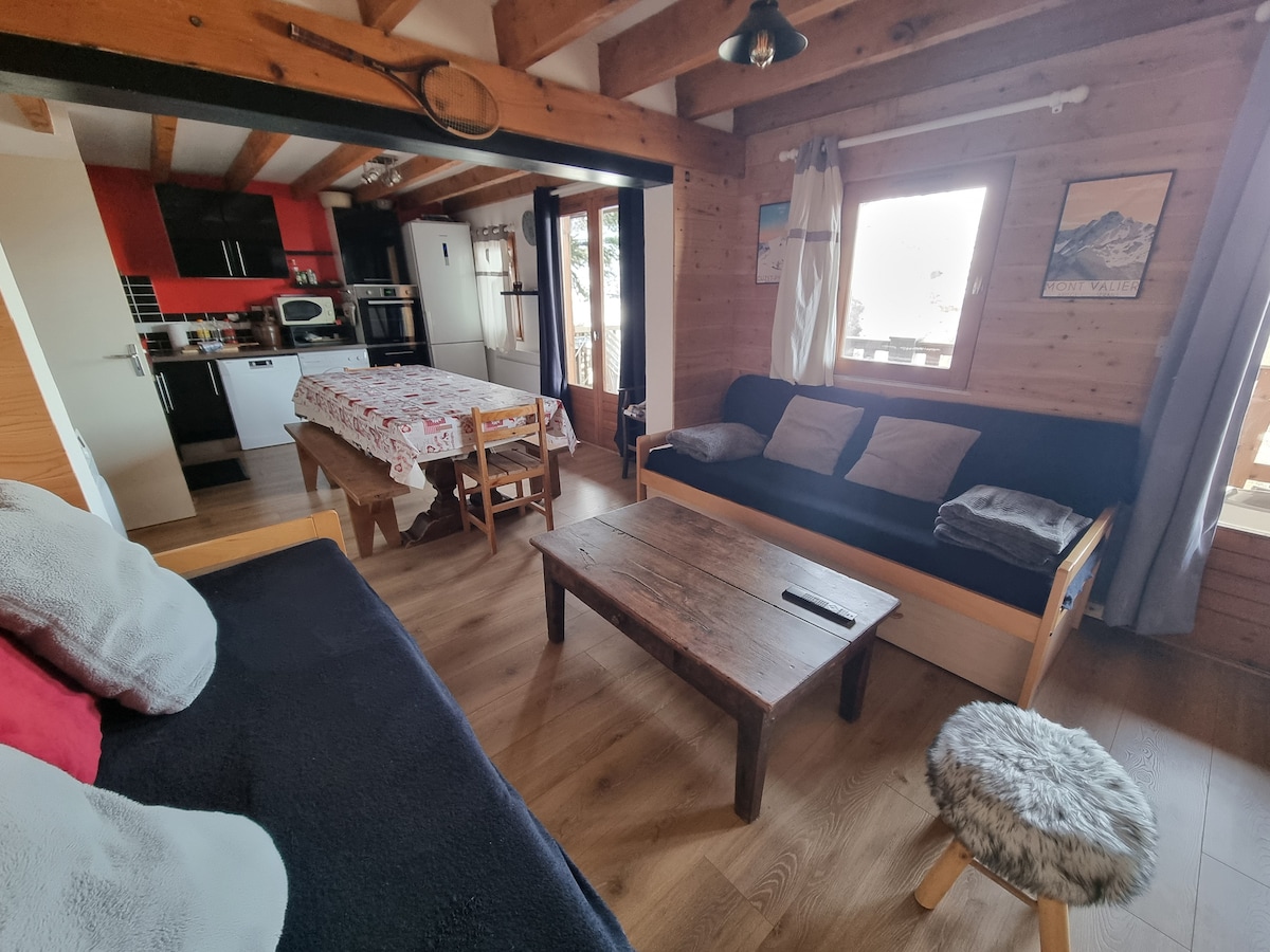 The spacious living area features wooden beams and a cozy seating arrangement with dark sofas. A large dining table is set for meals, while a well-equipped kitchen is visible in the background. Natural light floods in through windows, enhancing the warm wood tones throughout.