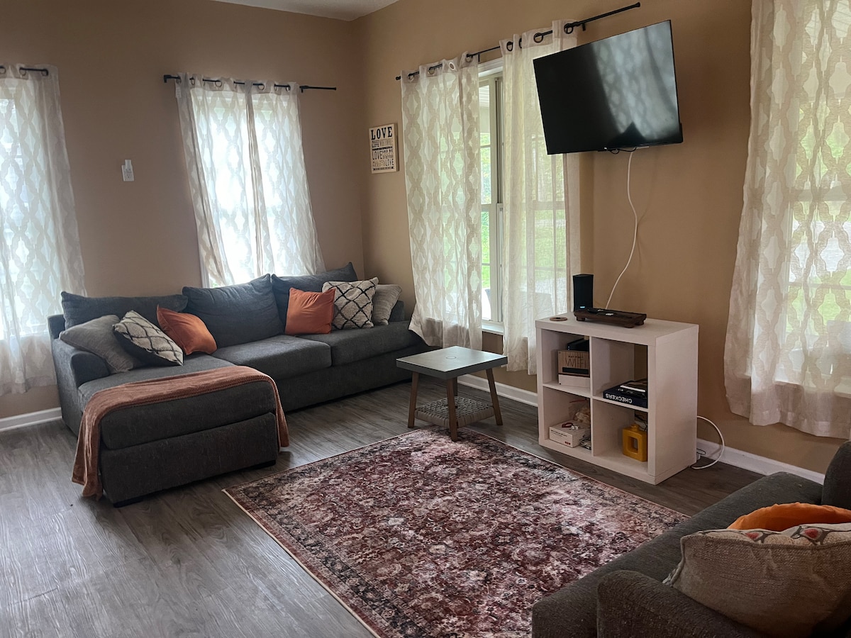 A welcoming living area features a large sectional sofa with decorative cushions and a matching armchair. A small coffee table sits on a patterned area rug, while a white media console displays speakers and a television. Natural light fills the space through two large windows dressed with light curtains.