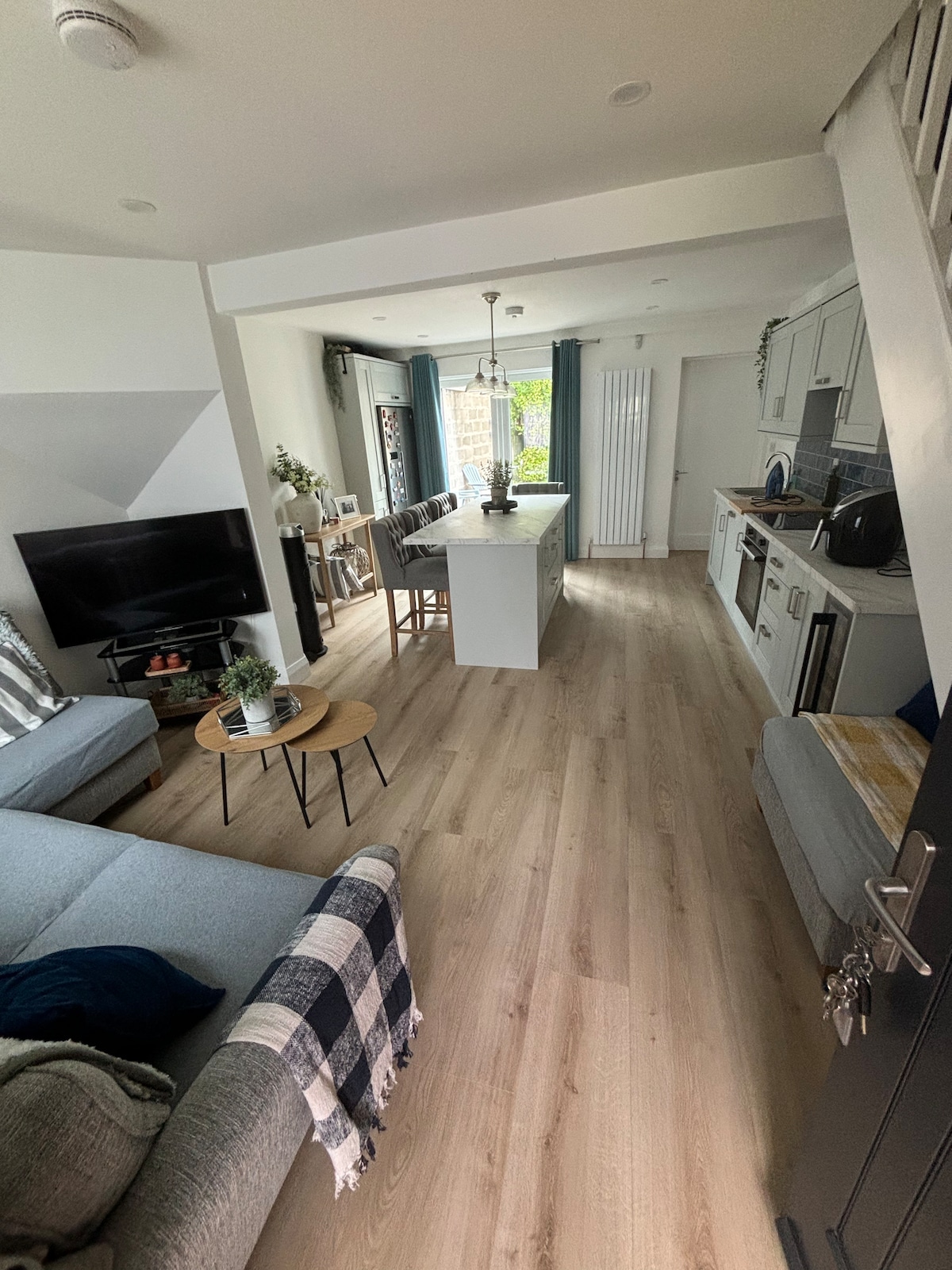 An inviting open-concept living space showcases a modern kitchen with a central island and seating. A comfortable sofa with a throw blanket is positioned near a television. Natural light fills the room through large windows, enhancing the wood-plank flooring and contemporary decor.
