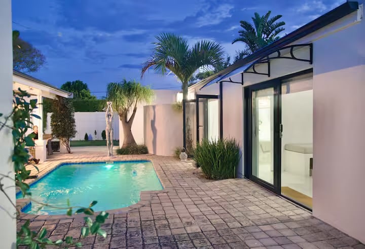 An outdoor area is highlighted by a tranquil pool surrounded by a stone-paved patio and lush greenery. Modern glass doors offer access to the interior, while palms and sculptural elements enhance the serene atmosphere under a twilight sky.