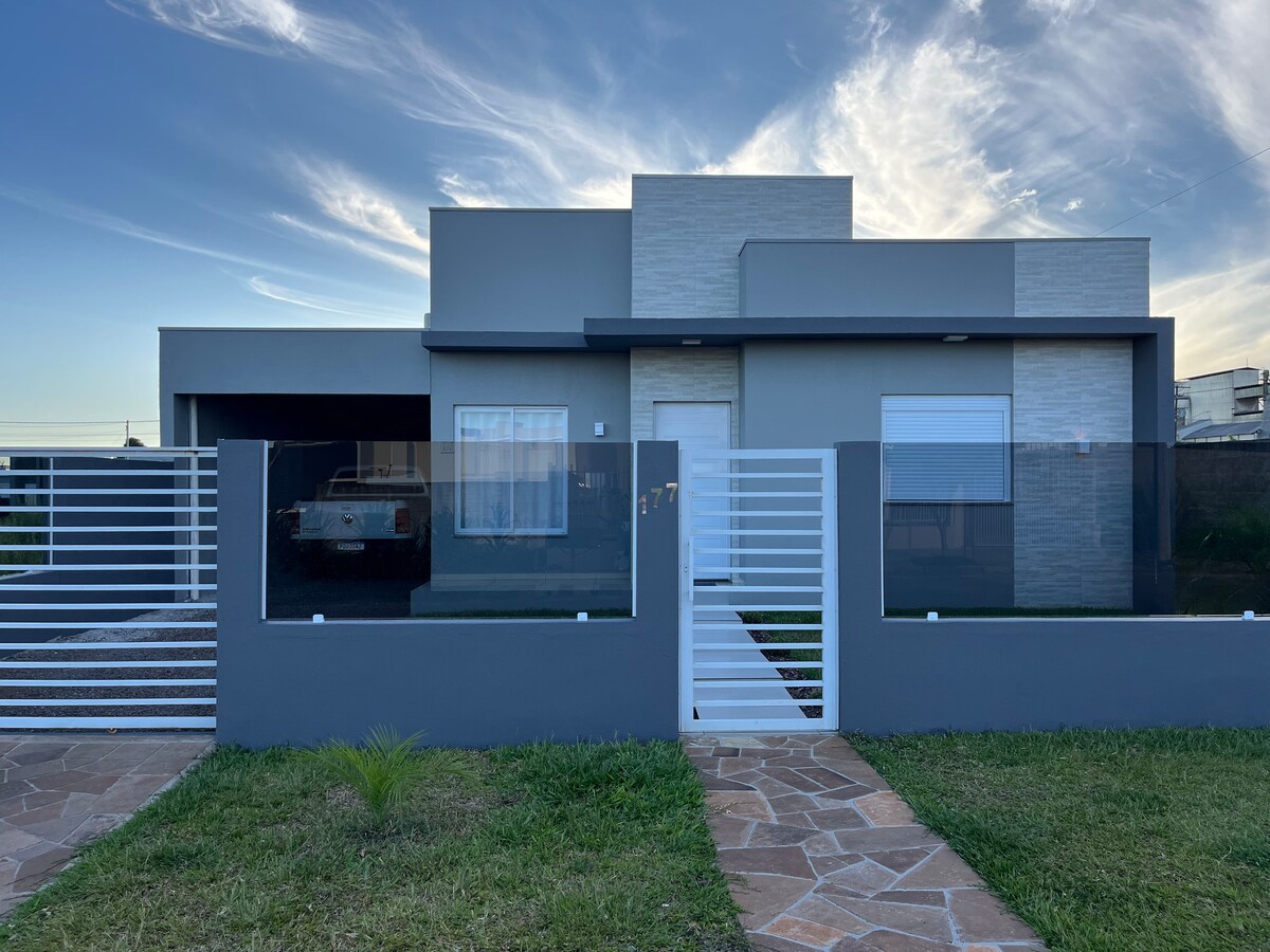 A modern single-story home with a light gray facade is framed by a low fence. The driveway features a gravel path leading to a gated entry. Large windows allow natural light to enter, and a landscaped area with green grass is visible in the foreground.