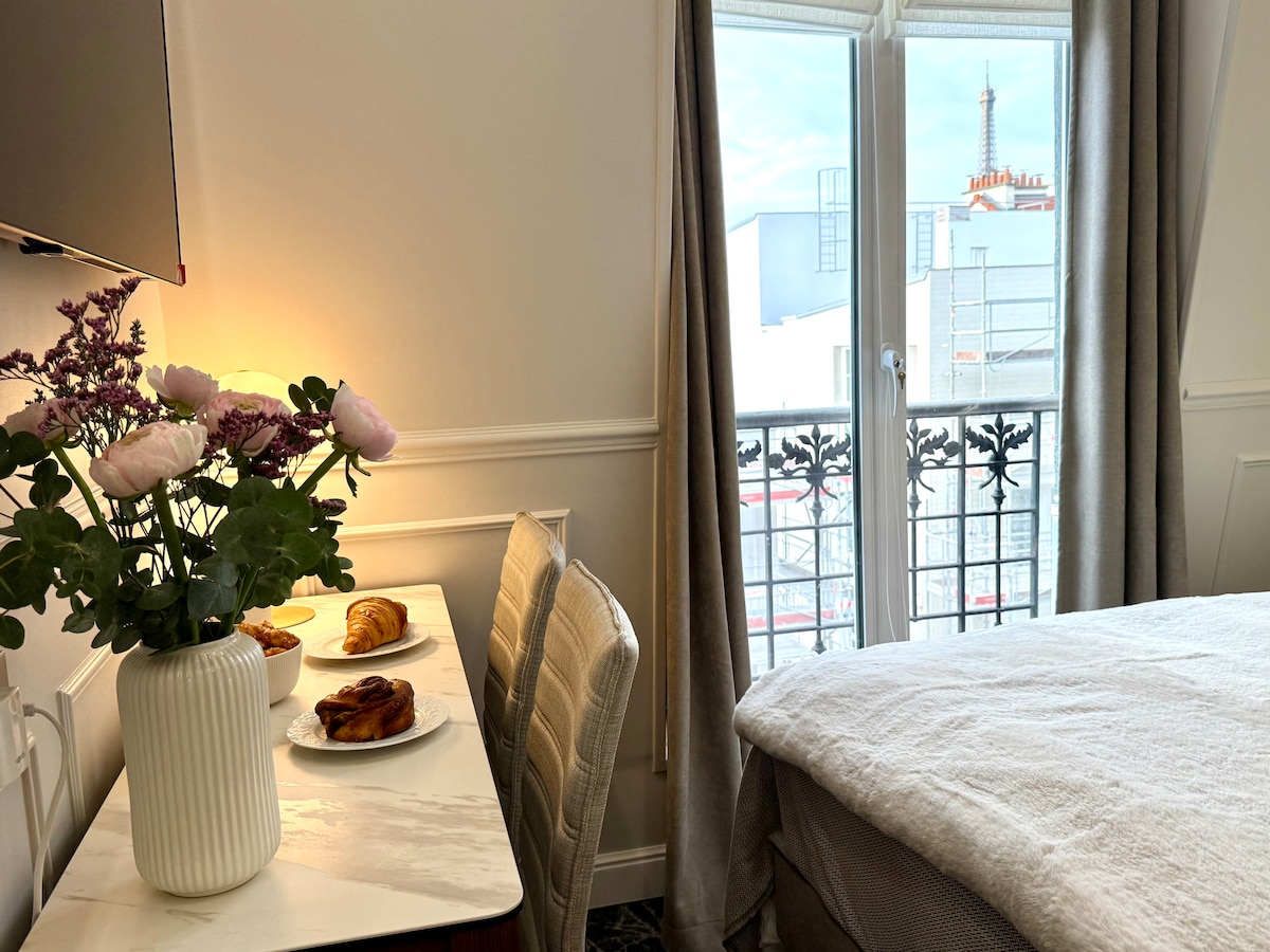 A cozy corner of the bedroom features a small table with a vase of flowers and pastries. A spacious bed is positioned nearby, and a large window offers a view of the Eiffel Tower, enhancing the room's ambiance with natural light.