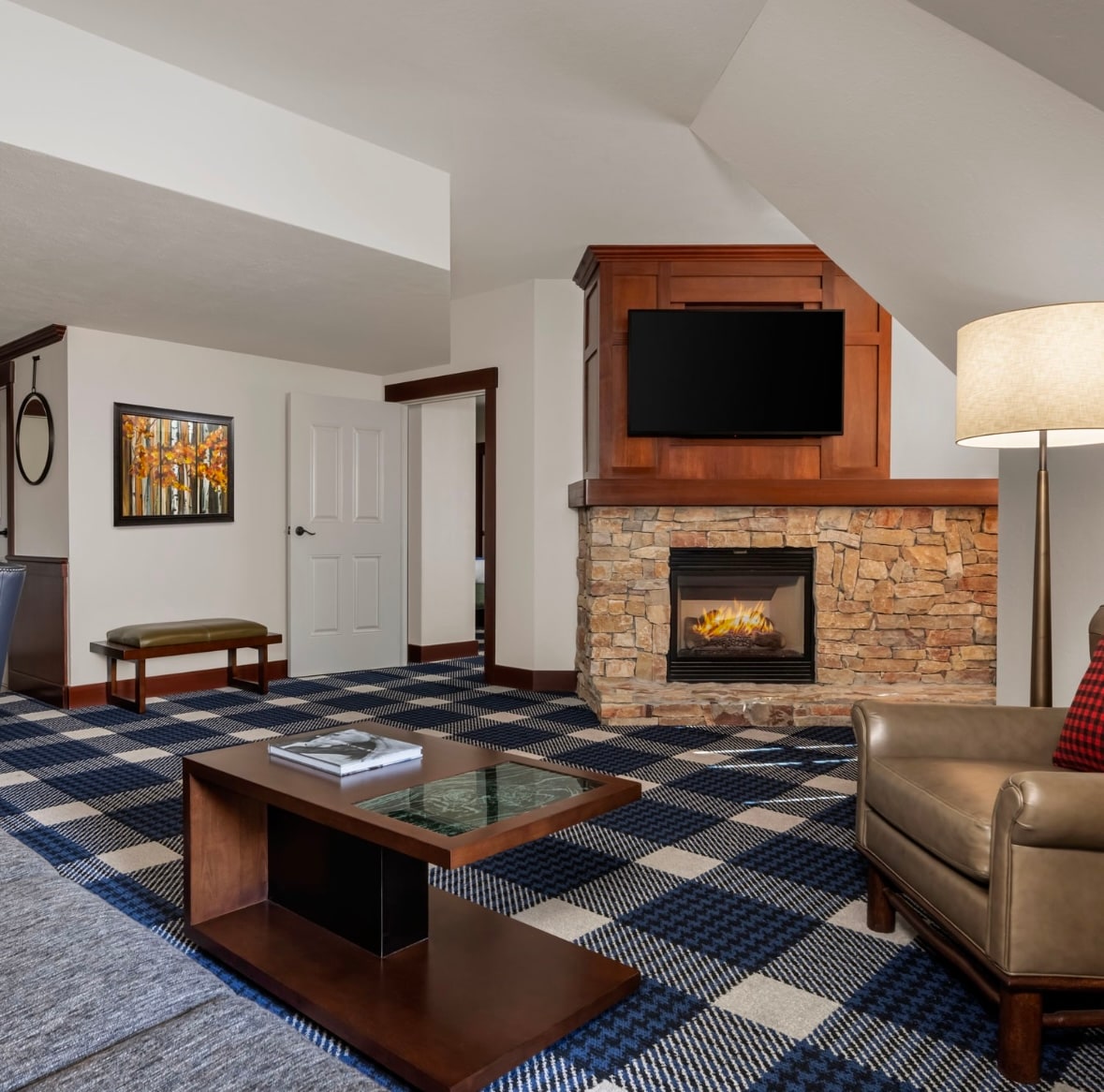 A stylish living area is depicted with a stone-clad fireplace and a flat-screen television mounted above it. A modern coffee table sits on a blue and white checkered carpet, complemented by a leather armchair and a floor lamp nearby.