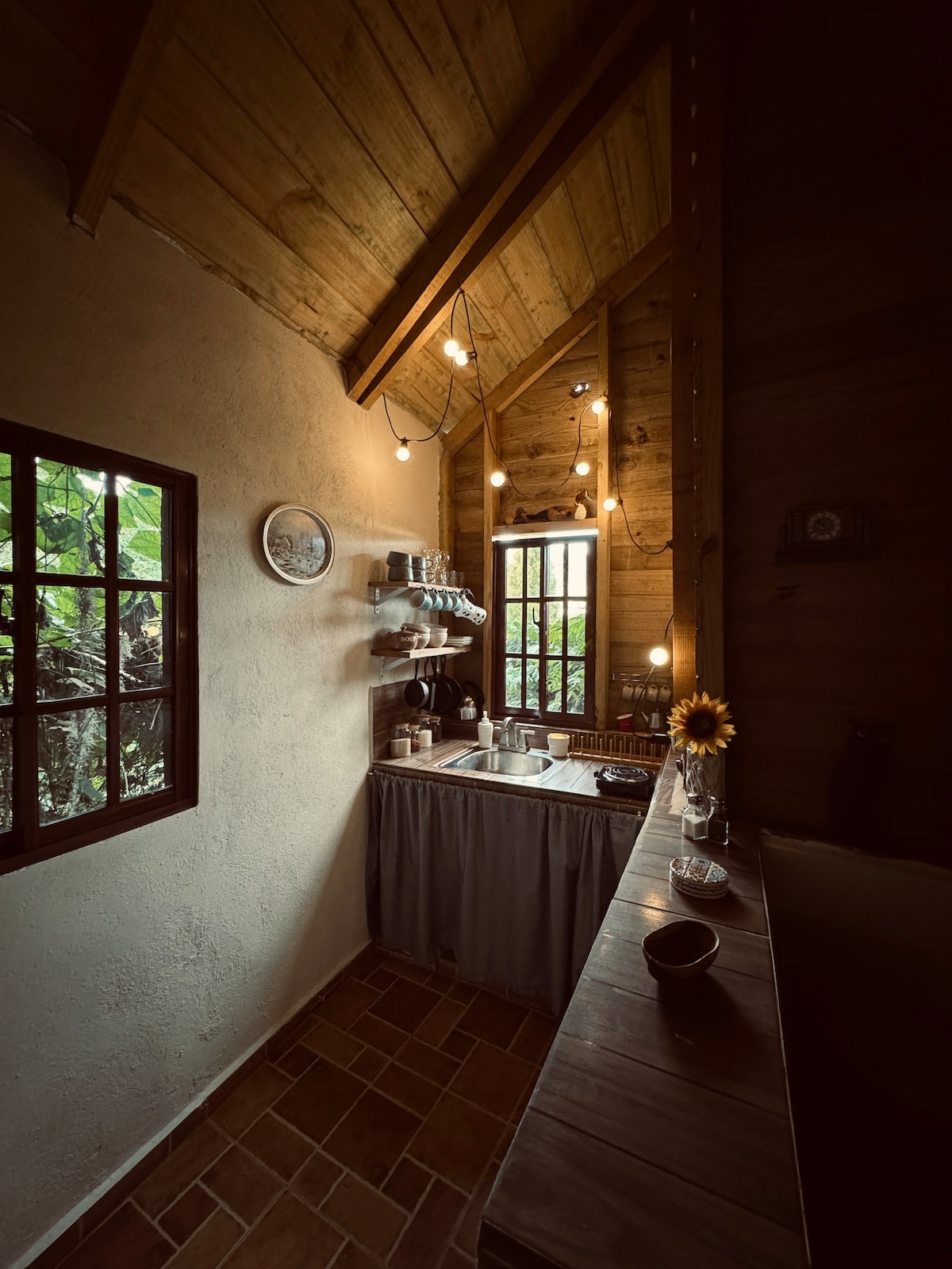A well-organized kitchenette features wooden ceilings with hanging light bulbs providing soft lighting. Stainless steel appliances are visible alongside a rustic countertop. A sink is located beneath a window, allowing natural light to filter in, and a sunflower in a vase adds a touch of color.