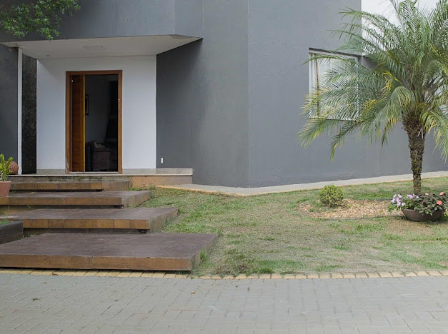 The entrance of the house is depicted, featuring a set of steps leading up to a door framed by a grey wall. Lush greenery, including a palm tree and flowerbeds, is visible, complementing the outdoor space and enhancing the overall welcoming appearance.