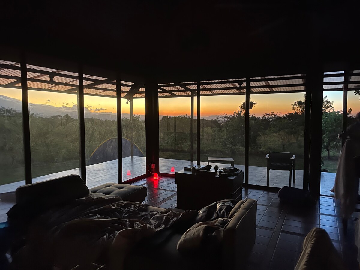 A spacious room features large glass doors that open up to a scenic view of the sunset, with colors reflecting off the landscape. Comfortable seating is arranged around a coffee table, and a soft blanket is draped over the bed, indicating a cozy atmosphere.
