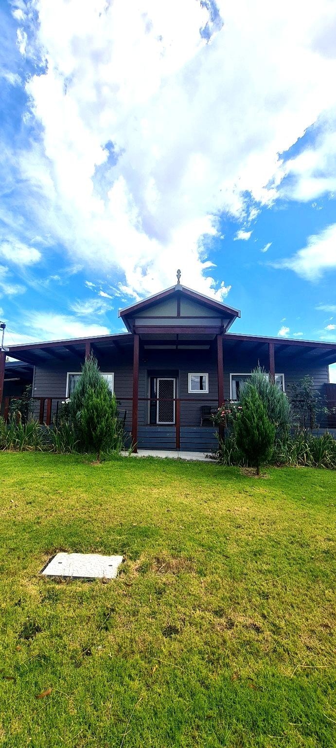 The exterior of a single-story home is portrayed, featuring a gabled roof and a central entrance. The house is surrounded by low greenery, and a grassy yard occupies the foreground. A clear blue sky with scattered clouds enhances the inviting appearance of the property.