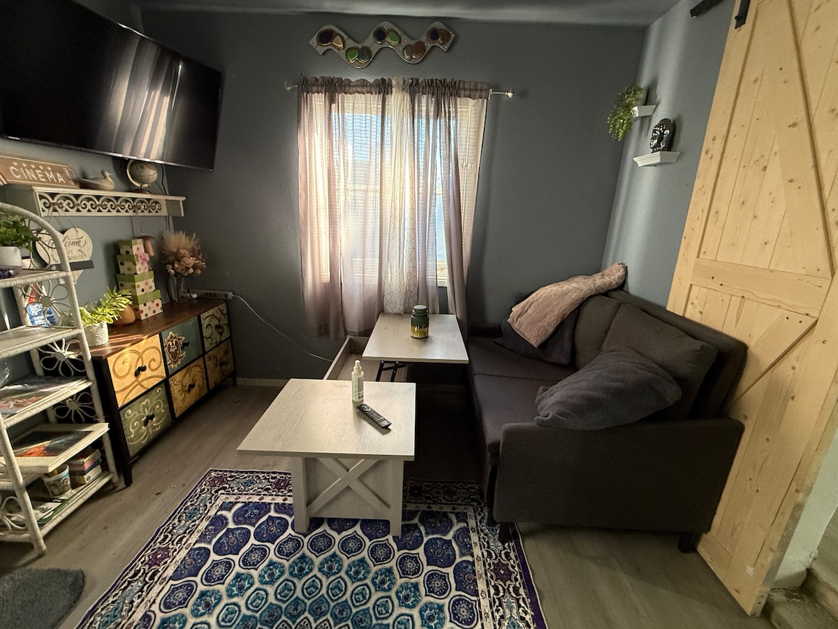 A cozy living area features a dark sectional sofa and a white coffee table. Natural light filters through sheer curtains, illuminating the space. Colorful storage boxes are displayed on a shelf, and a decorative rug adds warmth to the floor.