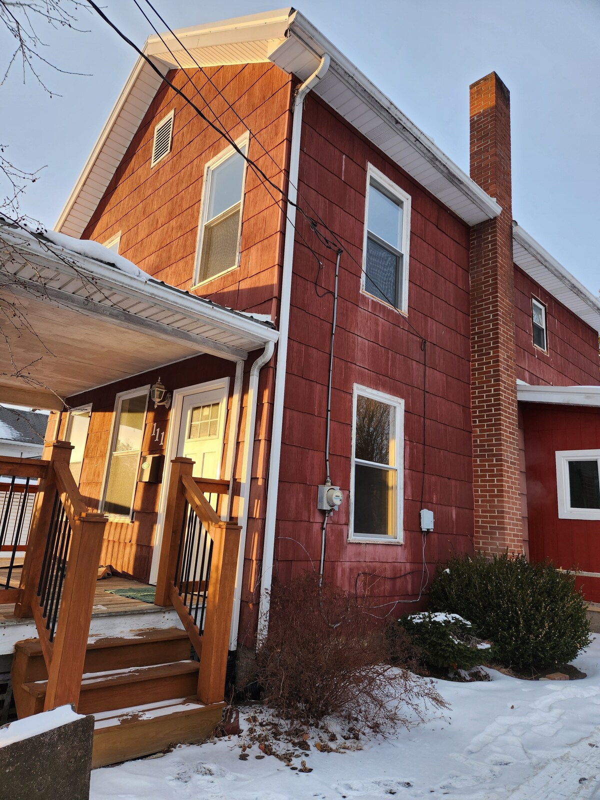 The exterior of the home is highlighted by red wood siding and features a covered porch with wooden steps and railing. A brick chimney extends from the side, while snow-covered ground adds a serene touch to the entrance.