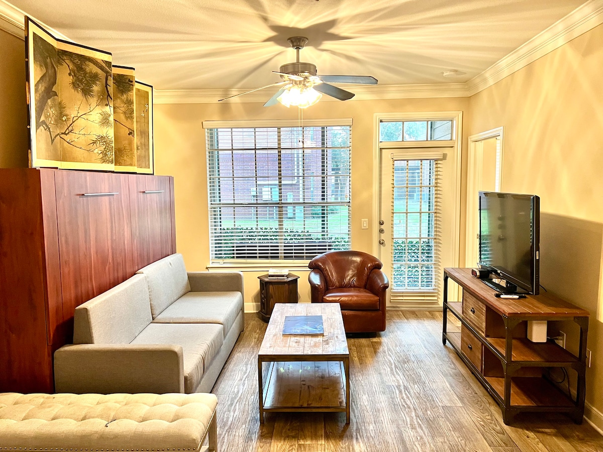 The living area features a comfortable sofa, a leather armchair, and a wooden coffee table. Natural light filters through large windows adorned with blinds, while a ceiling fan adds a touch of comfort. A television is positioned on a wooden media console against the wall.