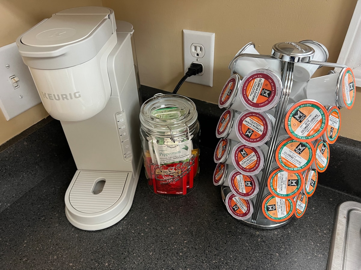 A modern coffee station is featured with a Keurig machine beside a glass jar filled with assorted tea bags. A carousel holding various K-cup pods is positioned next to the machine, offering a selection of flavors for guests.