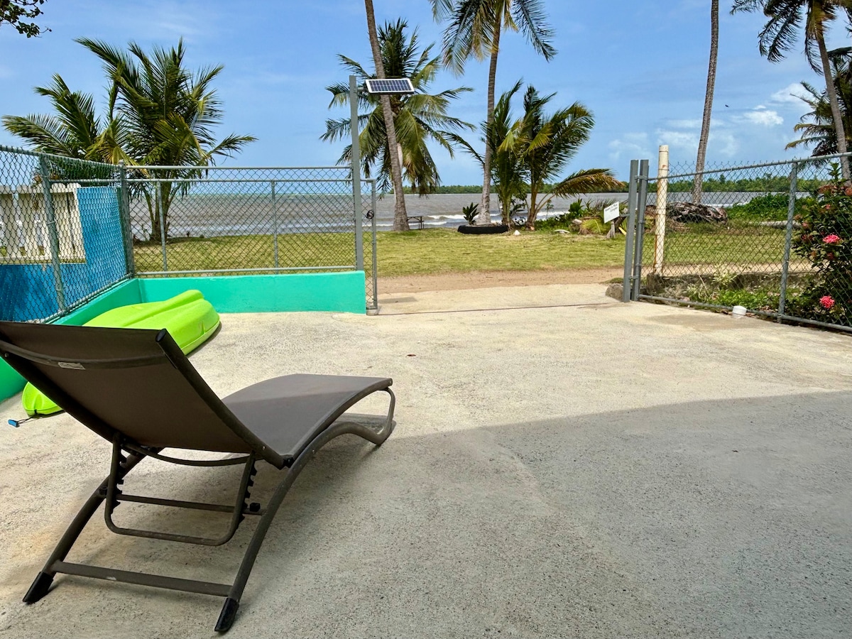 An outdoor area features a lounge chair positioned on a concrete surface, facing a green lawn and palm trees. A fence encloses the space, and a solar panel is mounted nearby. The view includes a glimpse of the coastline under a clear blue sky.