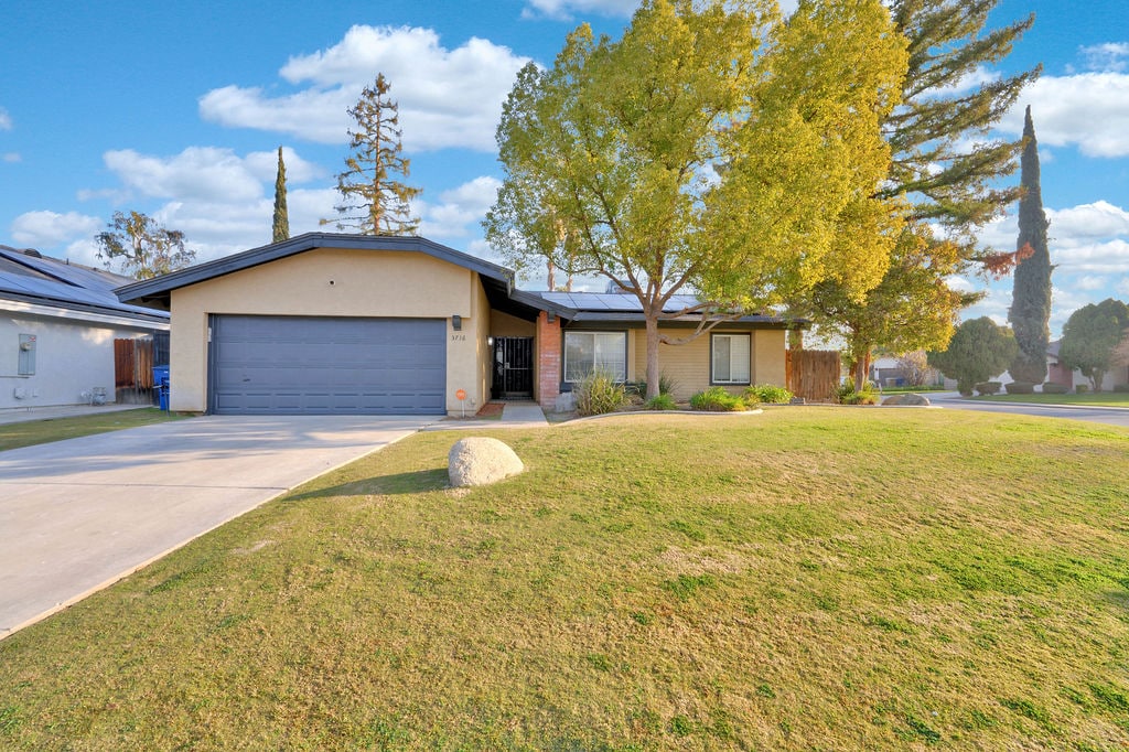 A single-story home is set amidst a well-maintained lawn, featuring a spacious driveway leading to a garage. Lush green trees surround the house, enhancing the outdoor space. The home’s entrance is framed by large windows that invite natural light into the interior.