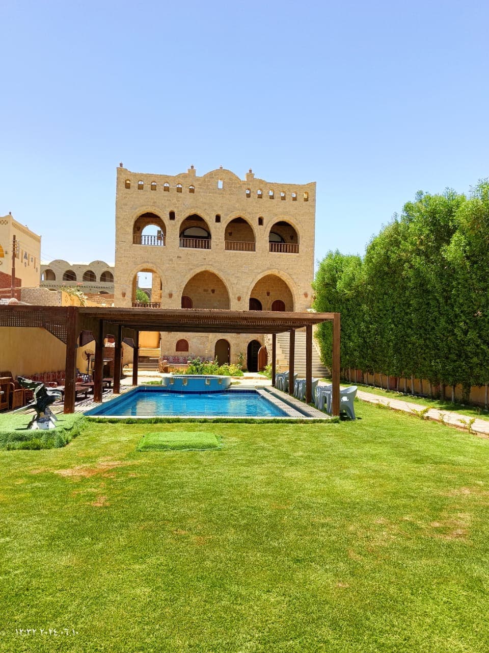 The outdoor area features a swimming pool surrounded by neatly trimmed grass and sun loungers. A large structure with arched windows rises in the background, complemented by lush greenery on either side, creating a serene environment for relaxation.