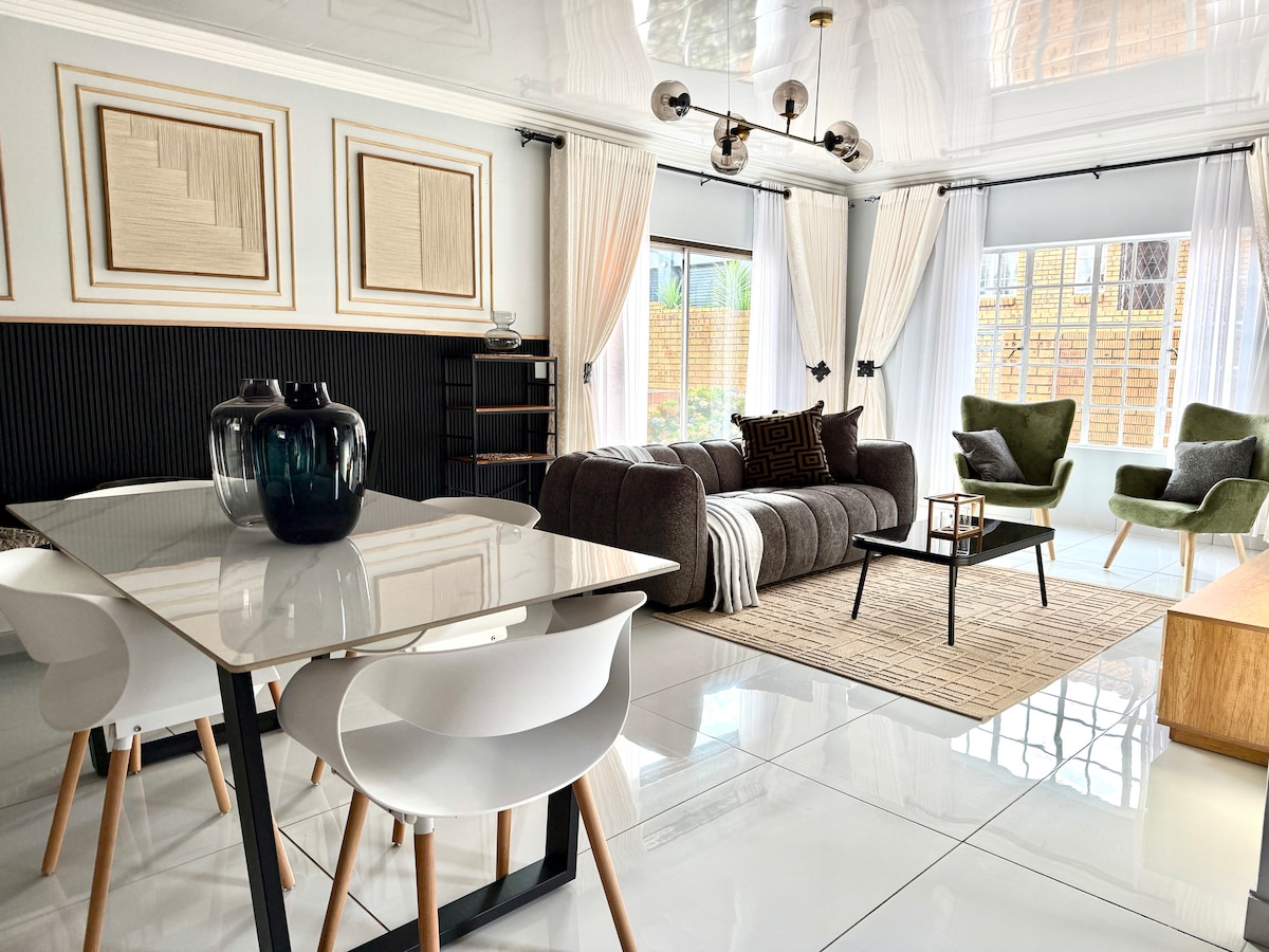 A modern living area is showcased with a sleek glass dining table and white chairs, complemented by a comfortable gray sofa and two green accent chairs. Natural light filters through large windows, illuminating the glossy floor and decorative wall art.