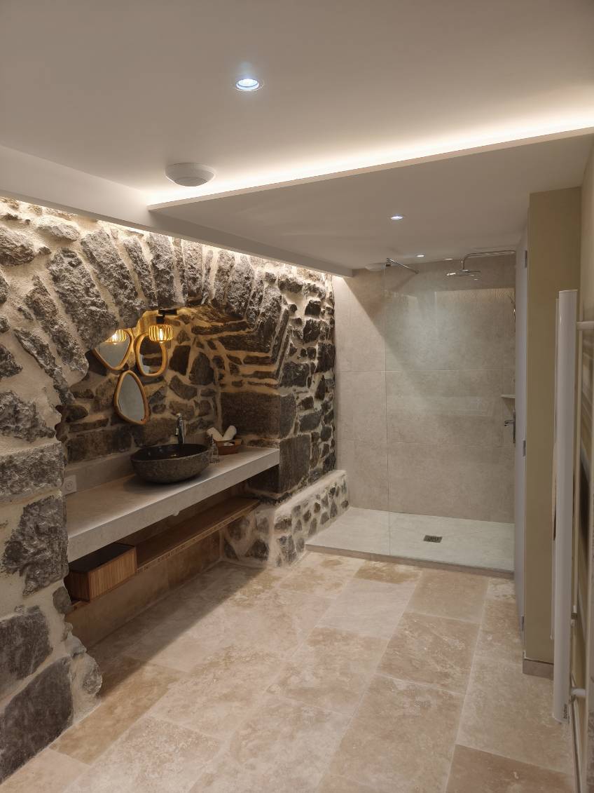 The bathroom features a natural stone wall with a built-in sink and a large mirror above. A spacious walk-in shower is designed with tiled walls. Soft lighting enhances the earthy tones of the stone and the serene ambiance of the space.