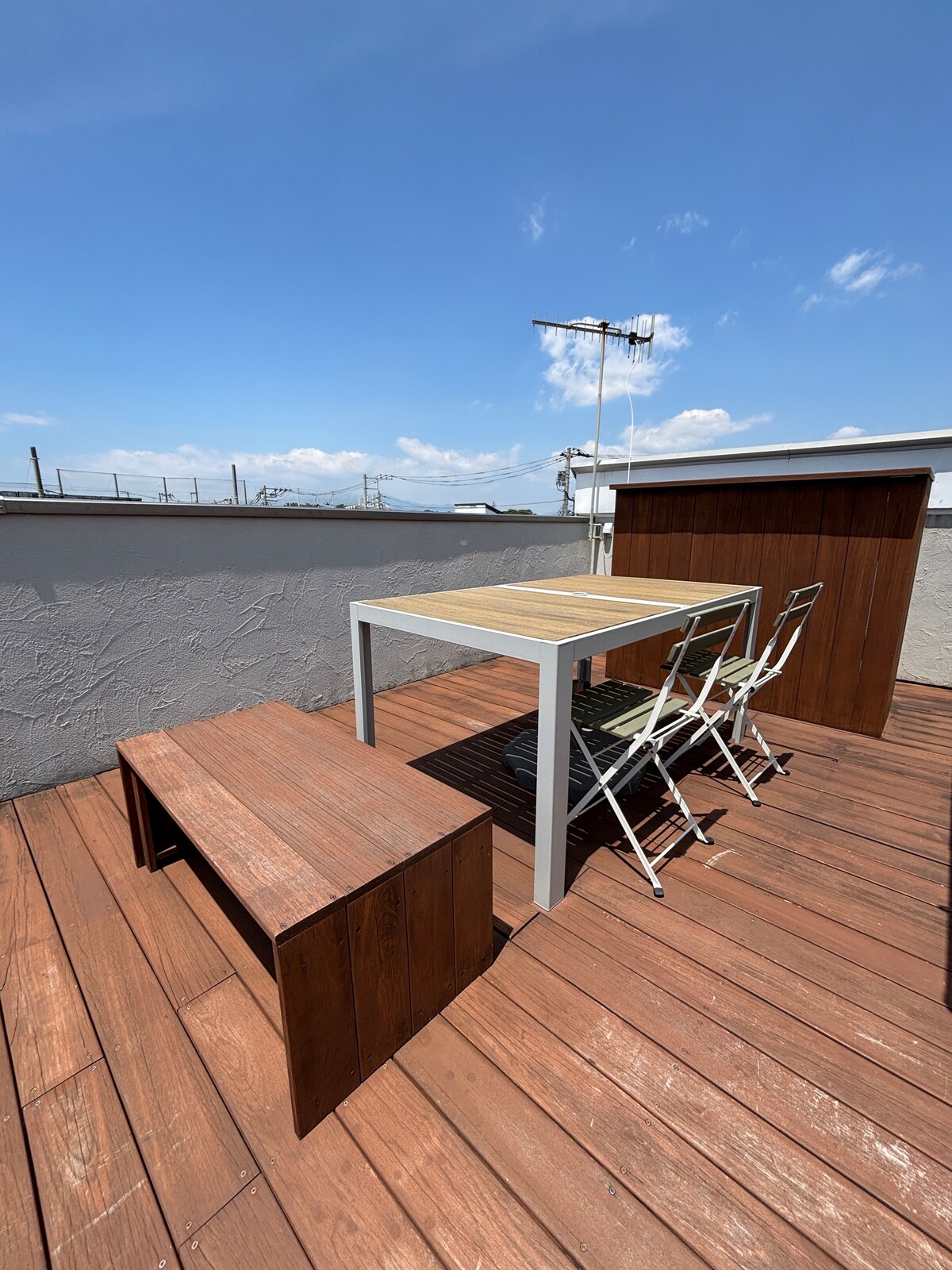 A rooftop deck is showcased with wooden flooring and a clear blue sky above. A rectangular table is accompanied by two folding chairs, and a wooden bench adds additional seating. The surrounding walls provide a sense of privacy.