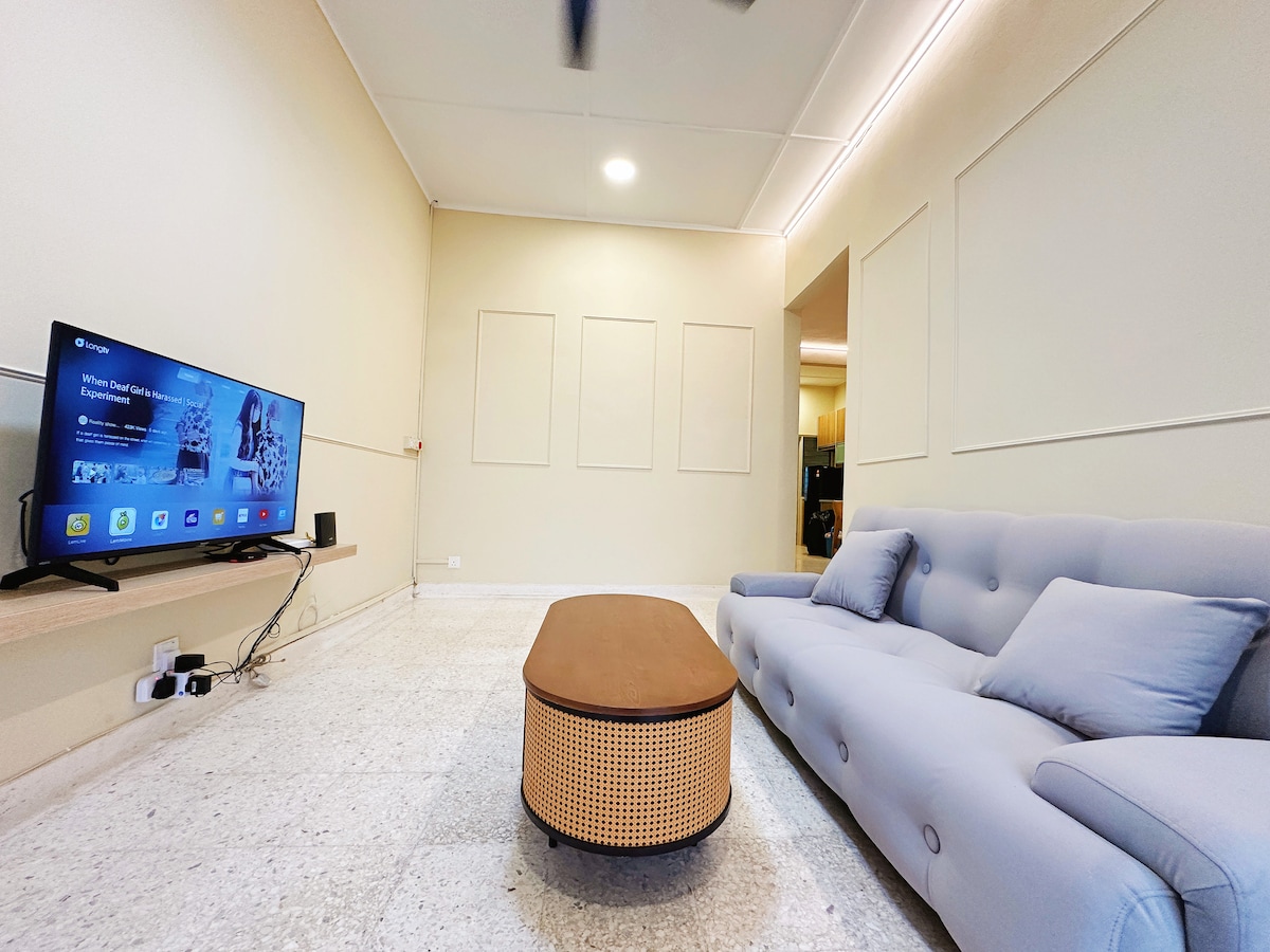 A bright living area features a modern couch with soft blue cushions. A low coffee table with a natural wicker design complements the space. A flat-screen TV is mounted on the wall, offering a contemporary entertainment option. The room is well-lit with soft overhead lighting.