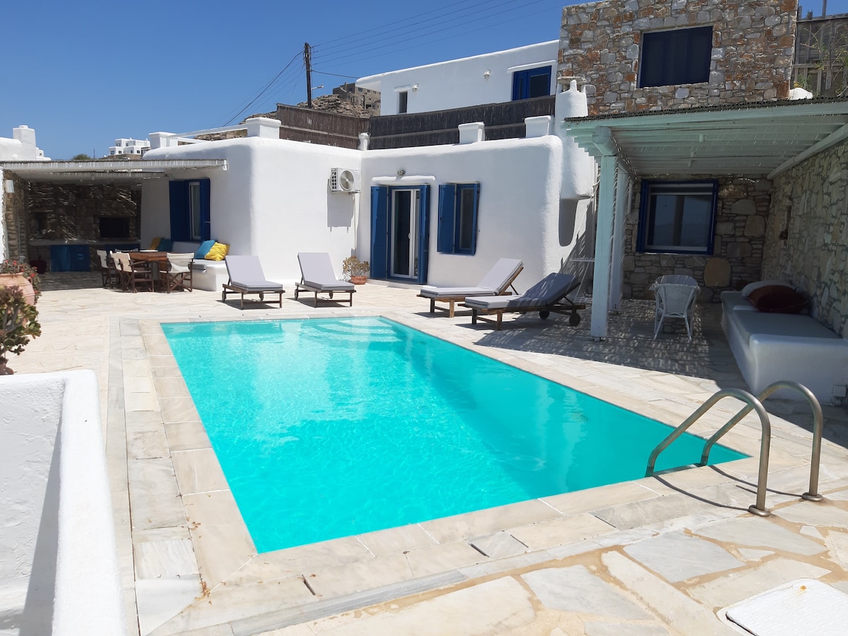 A private swimming pool is surrounded by a spacious terrace, featuring sun loungers for relaxation. The villa's white exterior contrasts with the blue windows, while nearby seating and dining areas are visible, inviting guests to enjoy outdoor meals.