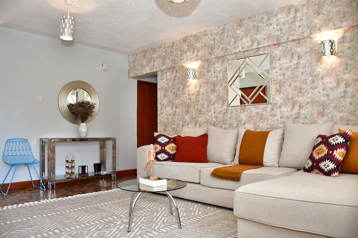 A comfortable living area is presented, featuring a spacious sectional sofa adorned with decorative pillows in various patterns. A round glass coffee table sits in front of the sofa, while a mirrored accent wall reflects the warm lighting from stylish wall fixtures. A blue chair adds a pop of color.