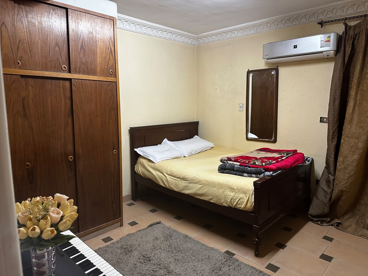 A modest bedroom features a wooden bed with a simple yellow bedspread and white pillows. A large wooden wardrobe stands against the wall, while a mirror is mounted nearby. A rug lies on the floor beside a keyboard, adding a touch of decor.