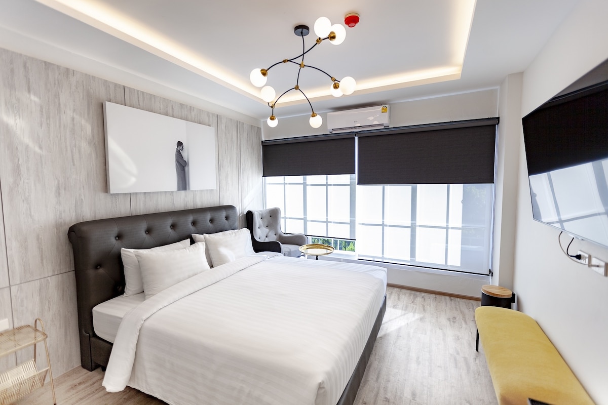 A spacious room features a comfortable bed with crisp white linens. Large windows with dark shades allow for natural light. A stylish armchair and a small side table are positioned beside the bed. A wall-mounted television is visible, enhancing the modern design.