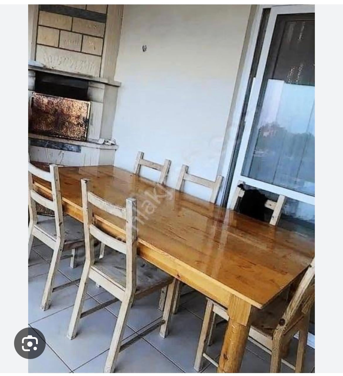 A wooden dining table is surrounded by six matching chairs, placed on a tiled outdoor space. A stone fireplace is visible in the background, and large glass doors allow natural light to brighten the area.