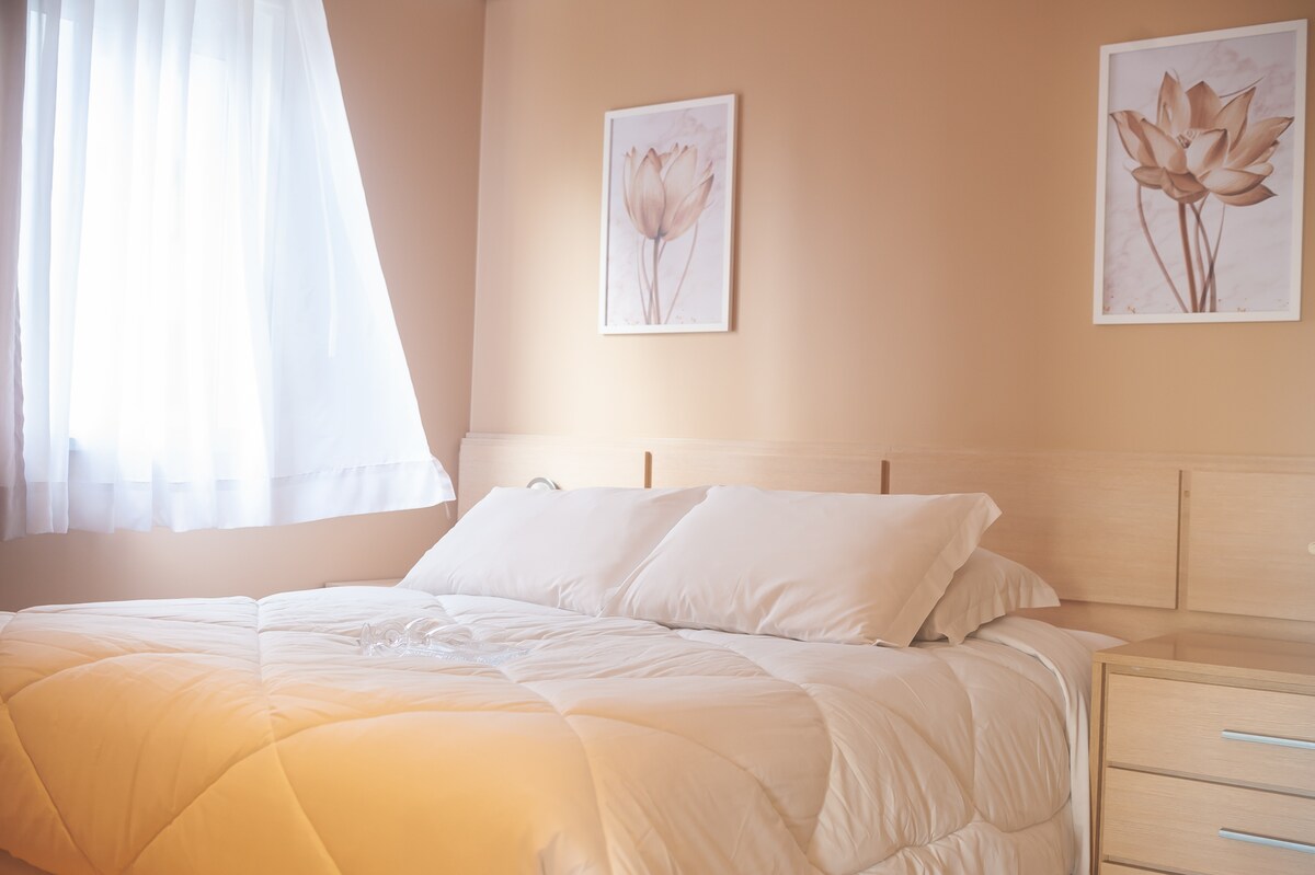 A bright and serene bedroom is displayed, featuring a spacious bed layered with a light-colored quilt. Soft natural light filters in through a large window with sheer curtains. Two floral artworks are mounted on the warm-toned walls, adding a touch of elegance.