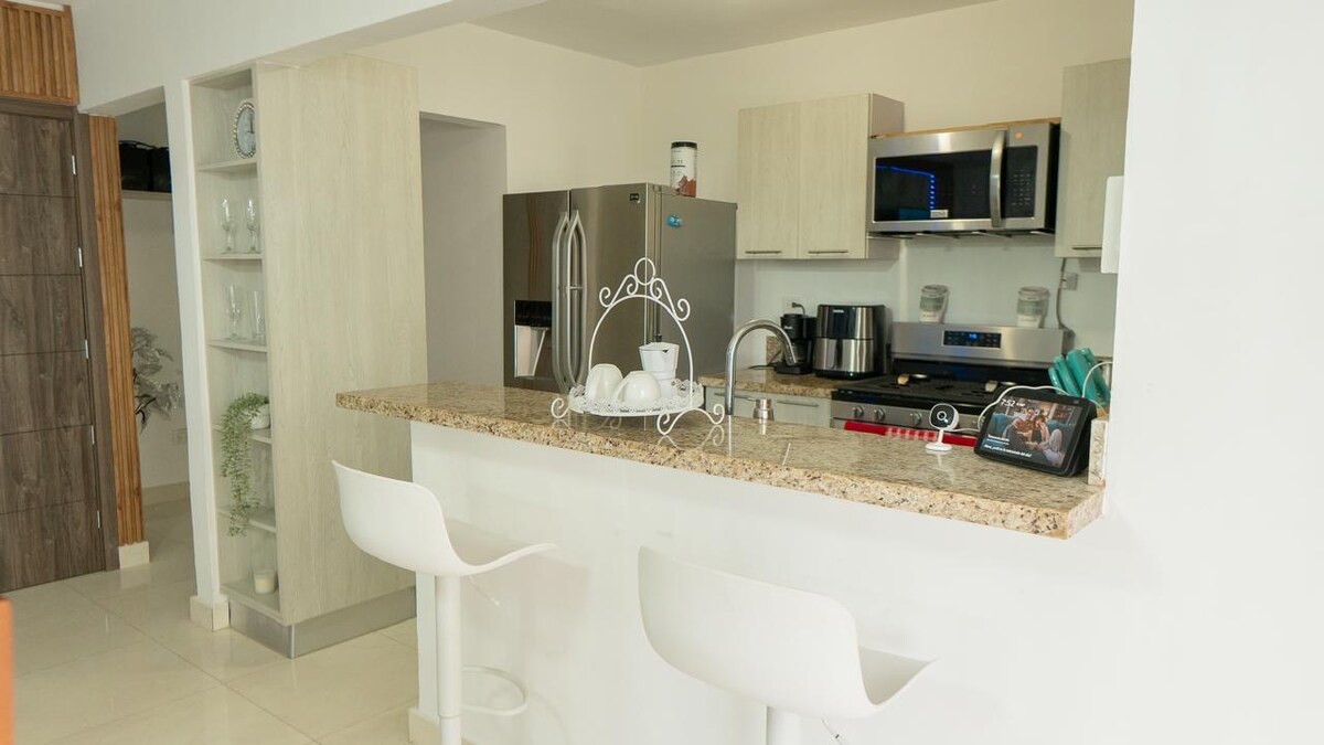 A modern kitchen space features an island counter with two bar stools. Stainless steel appliances, including a refrigerator and microwave, are visible. Light-colored cabinetry complements the granite countertop, which has additional kitchenware like a coffee maker and dishes set out.