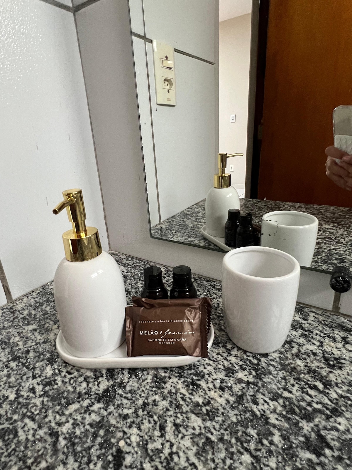 A white soap dispenser and a matching cup are placed on a granite countertop. Small dark bottles can be seen alongside a wrapped coffee treat. A mirror reflects the items, contributing to a functional and organized bathroom space.