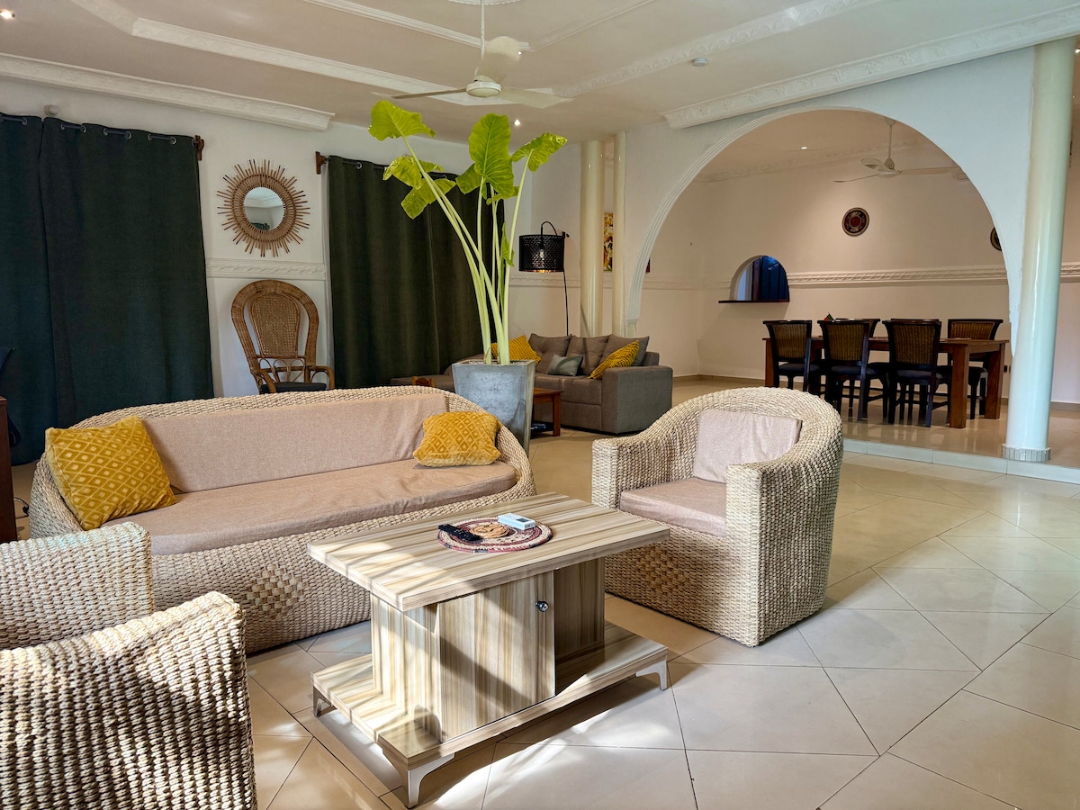 A spacious living area features comfortable seating arrangements, including woven furniture and vibrant cushions. Tall indoor plants add a touch of greenery, while dark curtains frame the windows. The open layout leads to a dining area, creating an inviting space for relaxation and socializing.