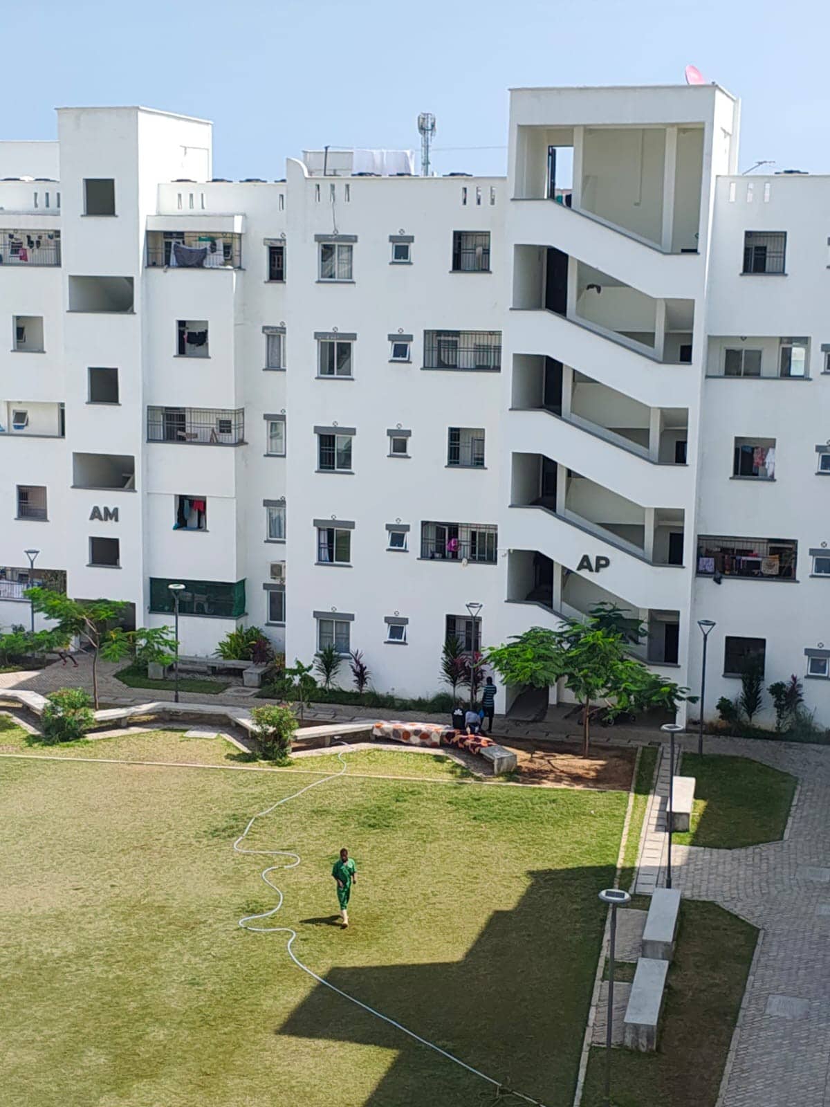 A multi-level building features a modern architectural design with white walls and large windows. A small green area is visible in the foreground, along with a pathway. A few trees and landscaped areas enhance the outdoor space, while individuals are seen enjoying the communal environment.