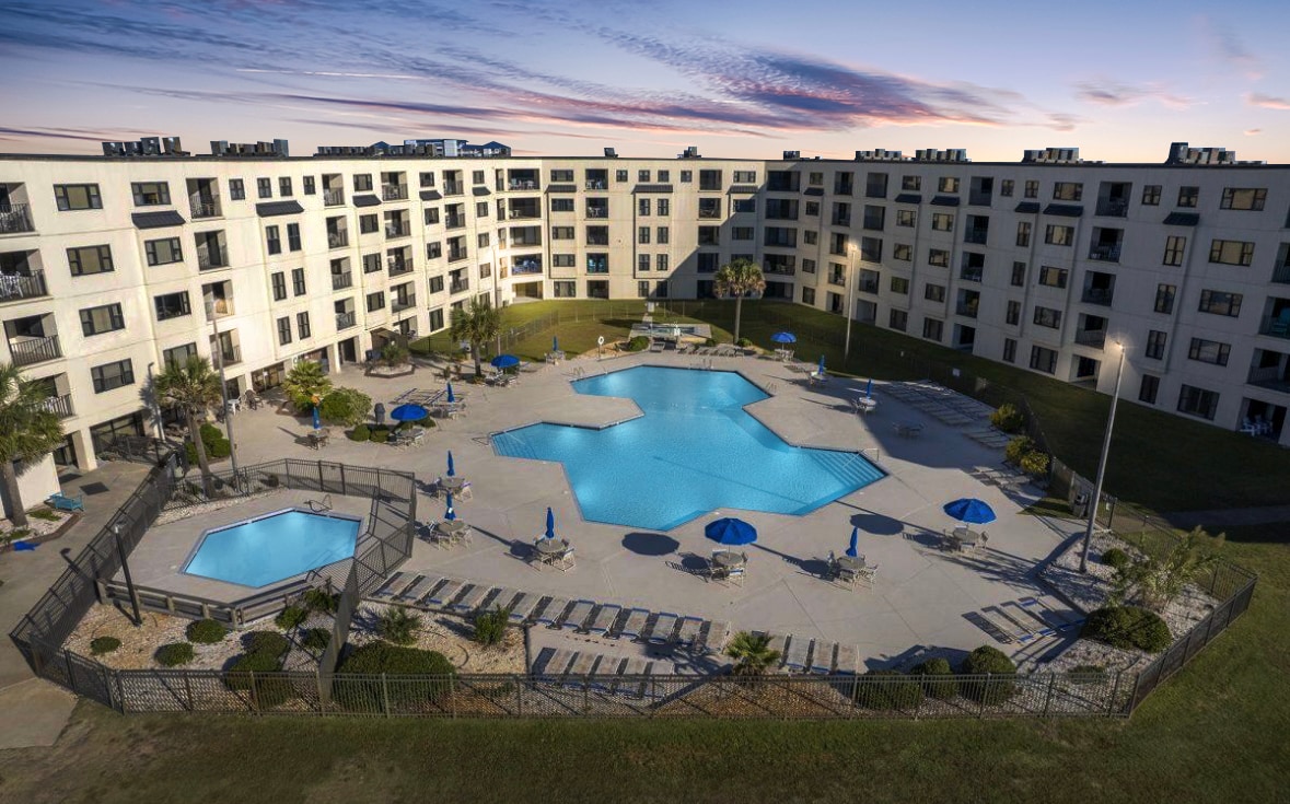 A central outdoor area is surrounded by a three-story condo building, featuring a large, shaped swimming pool and an octagonal pool. Several lounge chairs with blue umbrellas are arranged around the pool area, set against a scenic backdrop of the evening sky.