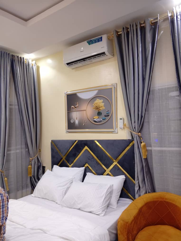 A comfortable sleeping area features a plush bed with white linens and an elegant headboard adorned with geometric patterns. Stylish grey curtains frame the window, and a modern air conditioning unit is positioned above, complementing the art piece showcased on the wall.