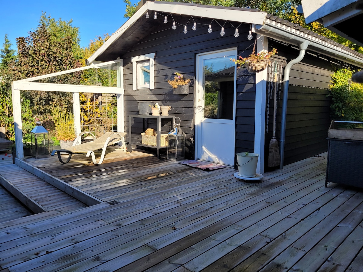A wooden deck extends in front of a dark cabin, featuring a covered seating area with a lounge chair. Decorative lights are strung along the roofline. A white door opens into the cabin, while planters and a cozy fireplace are visible nearby.