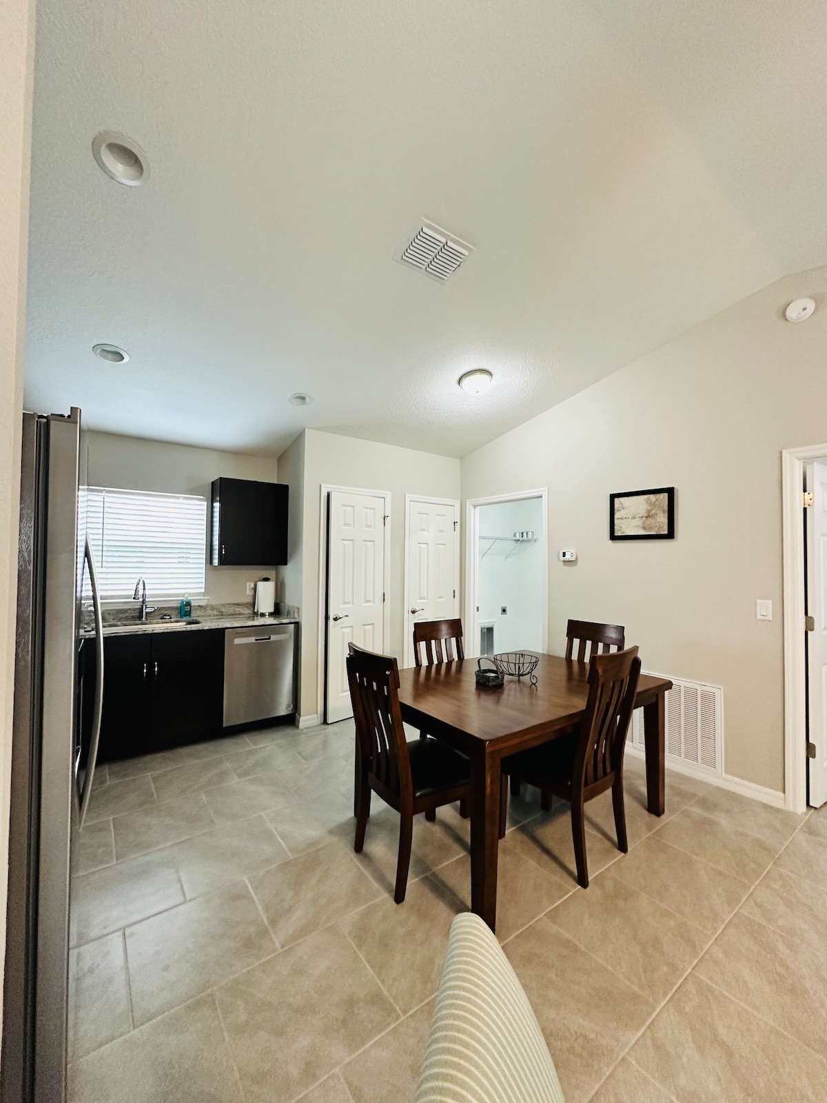 A modern kitchen area is visible, featuring stainless steel appliances and dark cabinetry. A wooden dining table is positioned centrally, surrounded by six chairs. Natural light flows in through the window, highlighting the light-colored tile flooring.