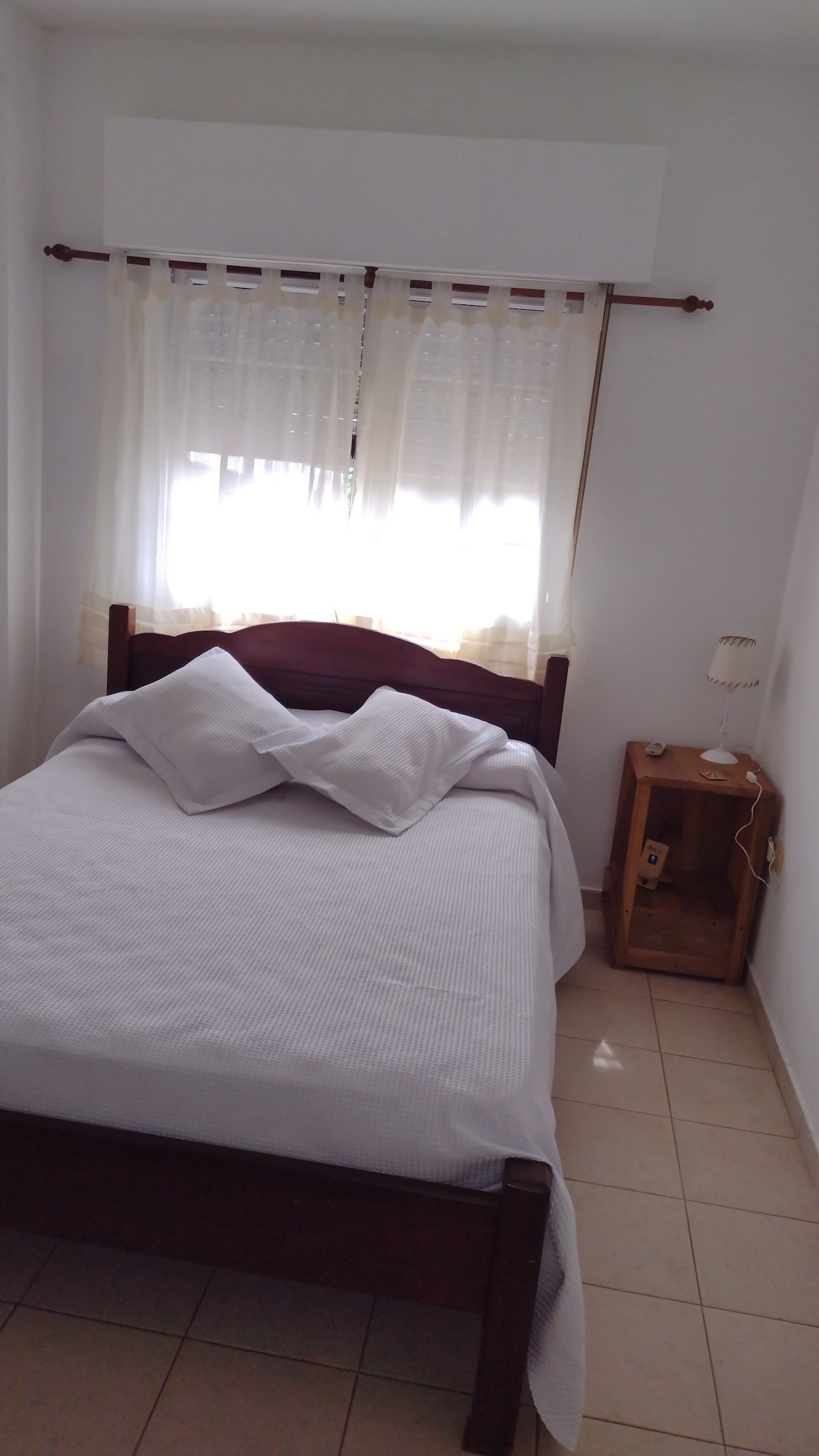 A well-lit bedroom features a single bed with neatly arranged pillows. Natural light filters through sheer curtains, illuminating the room. A bedside table holds a lamp, adding functionality to the space, while tiled flooring contributes to a clean and welcoming atmosphere.