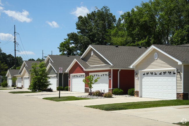 A row of single-story houses is presented, featuring various exterior colors including cream and red. Each house includes a spacious driveway and a well-maintained lawn, with trees and shrubbery enhancing the neighborhood's greenery. Clear blue skies and fluffy clouds add to the setting.